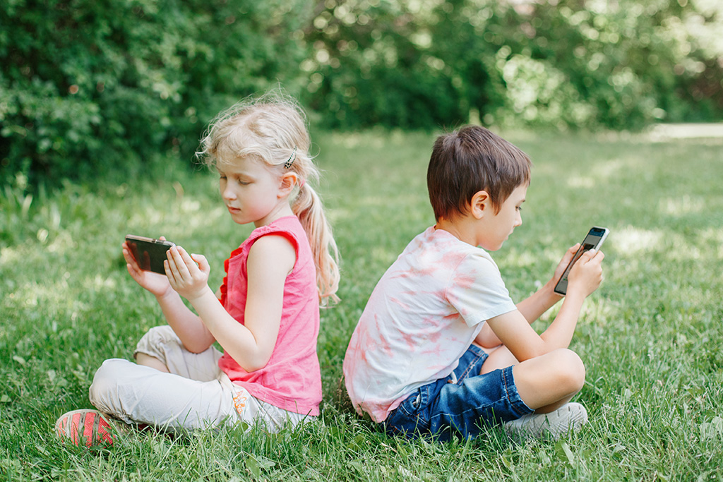 Two children sitting outside in the grass looking at mobile phones.